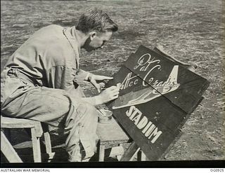 NADZAB, NEW GUINEA. C. 1944-02. FLIGHT SERGEANT F. "BLUE" ELLIS, CONCORD WEST, NSW, ONE OF THE FIRST AIR TRAINING CORPS CADETS IN OPERATIONS IN THE NEW GUINEA AREA, PAINTS A SIGNBOARD FOR THE ..