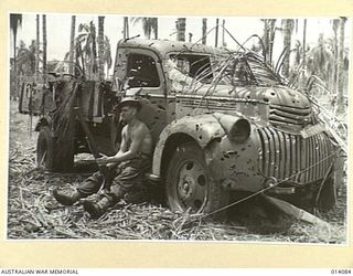 1943-01-14. PAPUA. GIROPI POINT. THIS IS WHAT CHEVROLET TRUCKS LOOK LIKE AFTER AUSTRALIAN MANNED TANKS HAD FINISHED WITH THEM DURING THE GIROPI POINT BATTLE. PTE. R. FRANCIS OF ADELAIDE HAS A REST ..