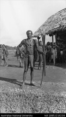 Man standing with bow and arrow with building and other individuals in background