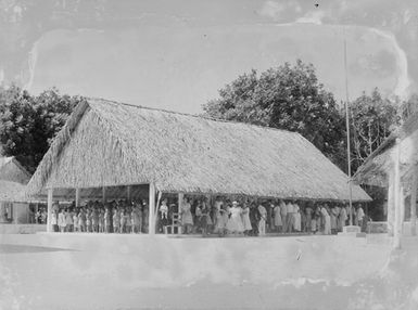 [A large group of people gathered underneath a building with thatch roofing]