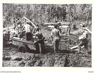 BOUGAINVILLE. 1945-07-21. TROOPS OF 3 SUPPLY DEPOT PLATOON, LOADING RATIONS ON TO A JEEP AND TRAILER AT THE OGORATA DETAIL ISSUING DEPOT. THE NATIVES IN THE BACKGROUND ARE LAYING CORDUROY IN AN ..