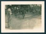 Two New Guinean women playing cricket with onlookers in background and one European man in foreground, Port Moresby, New Guinea, c1929 to 1932