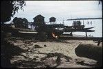 Manus: canoe-building, man heats pitch that has been applied to canoe, Pere village over-water houses in background