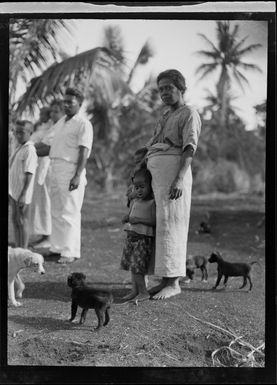 A portrait of a local Tongan mother and child with young dogs, with family members or neighbours behind, Tonga