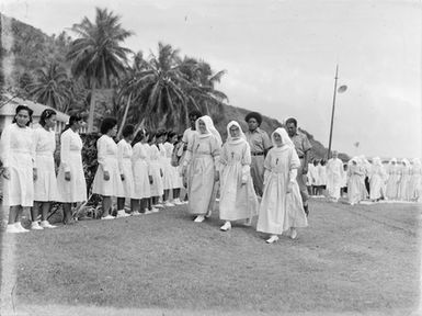 [Young pacific island women in uniform lined up watching a procession of missionaries]