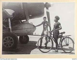 PIVA AIRSTRIP, JACQUINOT BAY, 1945-07-15. A NATIVE POLICE BOY WITH THE BICYCLE HE USES ON PATROL AROUND JACQUINOT BAY, LOOKING WITH INTEREST AT AN RNZAF CORSAIR ON THE TARMAC