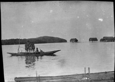 Four people standing and one seated in an outrigger canoe with marine houses in the background, Papua, ca. 1923 / Sarah Chinnery