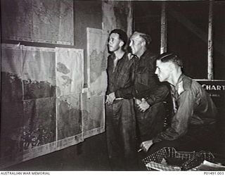 Probably MELBOURNE. C. 1944-09. MEMBERS OF THE MILITARY HISTORY SECTION STUDY MAPS OF NEW GUINEA IN THEIR OFFICE. LEFT TO RIGHT: JOHN CASTRAY, DRIVER J. W. ('SNOWY') STRANGE, SX2663 LIEUTENANT ..