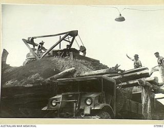 TOKO-BARARA ROAD, BOUGAINVILLE, SOLOMON ISLANDS. 1945-03-28. TROOPS OF THE 15TH FIELD COMPANY, ROYAL AUSTRALIAN ENGINEERS, USE A BULLDOZER TO FILL A TRUCK FROM A "CHINAMAN" CONTAINING TOP DRESSING ..