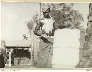 ALEXISHAFEN NORTH, NEW GUINEA. 1944-09-01. A NEW GUINEA NATIVE PAINTING A 44 GALLON DRUM WITYH WHITWASH AT THE CAMP OF THE 241ST SUPPLY DEPOT PLATOON