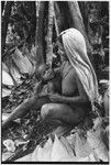 Pig festival, pig sacrifice, Tsembaga: woman with flattened leaves for sacrifice house thatching, holds infant