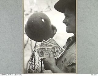 PAPUA. 1942-08-19. AN AUSTRALIAN SOLDIER EXAMINES A DAMAGED JAPANESE STEEL HELMET WHICH HAD BEEN PIERCED BY A BULLET FROM A THOMPSON SUBMACHINE GUN. HE ALSO HOLDS A HANDFUL OF JAPANESE BANKNOTES. ..