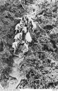 BULLDOG ROAD, NEW GUINEA. 1943-07-19. NATIVES CARRYING COMPRESSOR PARTS DOWN A SLIPPERY RAVINE AT ECCLESTONE GAP