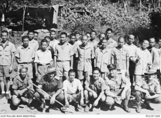 Rabaul, New Britain, 1945-10. Informal outdoors portrait of three Australian officers, members of a postwar recovery party, with a large group of Chinese prisoners-of-war (POWs) that they have ..