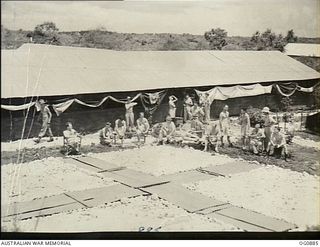 KIRIWINA, TROBRIAND ISLANDS, PAPUA. 1943-11-27. CONVALESCENT PATIENTS SITTING IN THE SUN AT NO. 3 MEDICAL RECEIVING STATION RAAF. THE BIG RED CROSS SET IN A WHITE CORAL BACKGROUND IS MADE OF ..