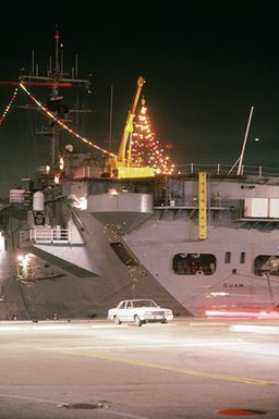 A port quarter view of the amphibious assault ship USS GUAM (LPH-9) decorated in Christmas lights for the holiday season. The ship is tied up at pier 4