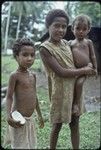 Girl holds a younger child, another girl holds split coconut, both of the younger children wear shell necklaces