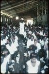 Group seated in a building, with Holy Mama in center; most are in white dress with red flowers in hair