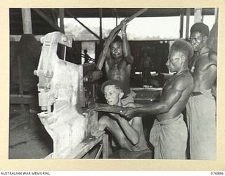 LAE AREA, NEW GUINEA. 1944-11-15. VX117885 CRAFTSMAN R.D. CURRAN, ASSISTED BY AUSTRALIAN NEW GUINEA ADMINISTRATIVE UNIT NATIVES, SHEARING HEAVY STEEL PLATE ON A GUILLOTINE IN THE GENERAL ..