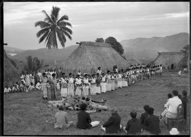 Fijian women dancing, with dwellings behind, a group in the foreground watching, and another group behind the women