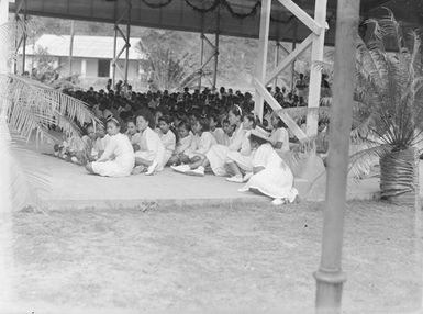 [Young pacific island children in uniform sitting inside an open air pavilion]