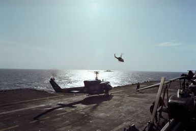 A UH-1 Iroquois helicopter is silhouetted by the sun as another UH-1 prepares to land on the flight deck of the amphibious assault ship USS GUAM (LPH 9). The ship is providing support to US Marines deployed in Lebanon as part of a multi-national peacekeeping force following confrontation between Israeli forces and the Palestine Liberation Organization