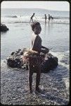 Girl in short fiber skirt walks on the fringing reef near Wawela village at low tide, other children fish on edge of coral reef in distance