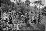Pig festival, stake-planting, Tuguma: decorated Tsembaga men wait for bamboo (on fire, center) to explode, signaling depature