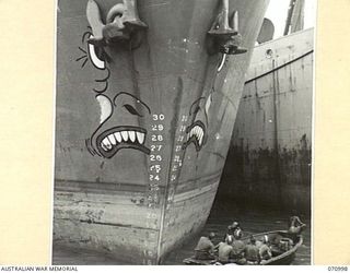 LAE, NEW GUINEA, 1944-03-09. SERVICEMEN EXAMINING THE GROTESQUE FACE ON THE BOW OF A LIBERTY SHIP UNLOADING AT THE WHARF