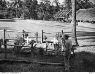 HIGITURA, NEW GUINEA. 1944-04-27. MR J. R. HALLIGAN, SECRETARY, DEPARTMENT OF EXTERNAL TERRITORIES (1), STANDS ALONGSIDE A SMALL PLOT IN THE SANGARA MISSION GROUNDS CONTAINING THE GRAVES OF MAY ..