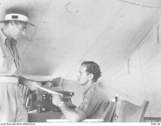 WEWAK POINT, NEW GUINEA. 1945-10-23. PRIVATE L. HARMES, 19 BRIGADE (1) HANDING THE REQUEST NUMBERS THAT BRIGADE PERSONNEL WISH TO HAVE RELAYED OVER THE AIR TO SERGEANT N. BRAY (2), AT RADIO STATION ..
