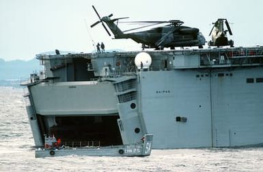 An LCM-6 mechanized landing craft is piloted past the docking well of the amphibious assault ship USS SAIPAN (LHA-2) during NATO Exercise Northern Wedding '86. A CH-53E Super Stallion helicopter and a CH-46 Chinook helicopter are on the flight deck