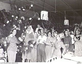 RABAUL, NEW BRITAIN, 1945-12-20. DURING THE CHRISTMAS PARTY HELD IN WARD 8, 118TH AUSTRALIAN GENERAL HOSPITAL, CAPTAIN V. H. HEGARTY, DRESSED IN THE UNIFORM OF A NURSING SISTER, PLAYED THE PART OF ..