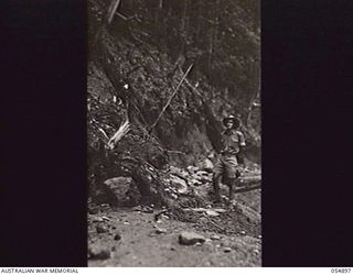 BULLDOG-WAU ROAD, NEW GUINEA, 1943-07-15. LIEUTENANT C. A. CASSON OF HEADQUARTERS, ROYAL AUSTRALIAN ENGINEERS, 11TH AUSTRALIAN DIVISION, INSPECTING A LANDSLIDE ON THE MOUNTAIN BENCHING AT THE 28 ..