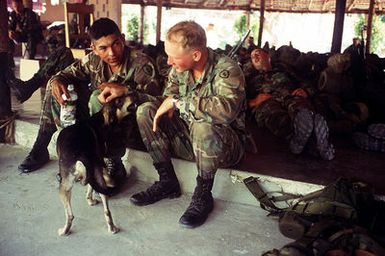 SPECIALIST Daniel Gonzales (left) and SPECIALIST David Cowan, both of Alpha Company, 2nd Platoon, 4th Battalion, 22nd Infantry, 2nd Brigade, 25th Infantry Division (light), Schofield Barracks, Hawaii, sit on a curb to pet a dog at the Special Forces compound in Mirebalais, Haiti in the late afternoon of 21 February 1995. Other soldiers can be seen in the background resting and sleeping. They will stay overnight at the compound before proceeding to Loscahobas, Haiti, where they will conduct foot patrols and set up vehicle checkpoints during Operation Uphold Democracy