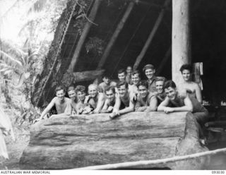 BOUGAINVILLE, 1945-06-07. MEMBERS OF 2/8 COMMANDO SQUADRON LEANING ON A NATIVE DRUM, A HOLLOWED LOG, OUTSIDE THE KARAMUT (SING-SING) HOUSE AT B TROOP HEADQUARTERS, NAIRONA. THE VILLAGE WAS ..