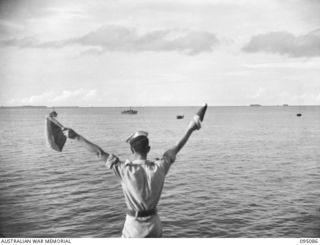 AT SEA, OFF BUIN, BOUGAINVILLE. 1945-08-20. A SIGNALLER USING SEMAPHORE FROM THE BRIDGE OF THE CORVETTE HMAS LITHGOW, AS THREE RAN MOTOR LAUNCHES ESCORTING JAPANESE BARGES APPROACH CARRYING ENVOY ..