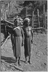 Men wear headdresses and shell valuables, stand near fence and ditch