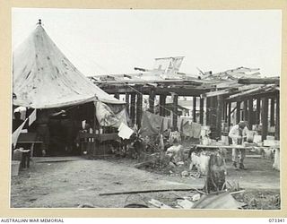 MADANG, NEW GUINEA. 1944-05-18. A PATIENT (1) CUTS BREAD FOR THE MID-DAY MEAL ALONGSIDE TENT WARD NO. 4 AT THE 111TH CASUALTY CLEARING STATION. THE RED CROSS FLAG FLIES BESIDE THE UNION JACK AT THE ..