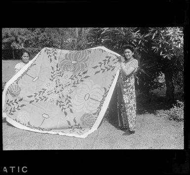 Two unidentified local woman displaying a tivaevae in front of trees, Rarotonga, Cook Islands