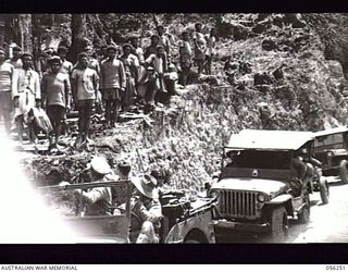 REINHOLD HIGHWAY, NEW GUINEA. 1943-08-25. SMILING NATIVES, WHO WORKED ON THE HIGHWAY, LINE UP NEAR FOX'S SADDLE TO GREET THE FIRST CONVOY TO TRAVERSE THE NEW ROAD. TRANSPORT AND TROOPS ARE FROM ..