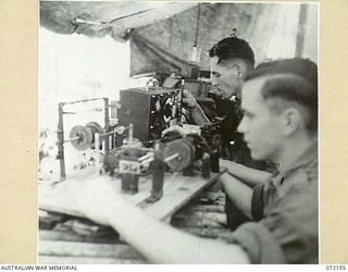 KILIGIA, NEW GUINEA. 1944-04-08. IN THE FOREGROUND QX52477 SIGNALMAN J. MCINTYRE (1), WINDS A TRANSFORMER COIL ON AN IMPROVISED WINDER WHILE IN THE BACKGROUND QX59856 CORPORAL E. NEILSON (2) ALIGNS ..