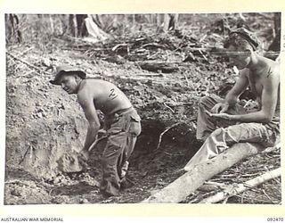 BOUGAINVILLE. 1945-05-22. PRIVATE R.W. MALSON, 24 INFANTRY BATTALION (1), SITTING ON A PIT EDGE ROLLING A SMOKE, WHILE PRIVATE F. NEAL (2), TAKES HIS TURN AT DIGGING A SLEEPING PIT ON EGAN'S RIDGE ..