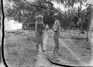 [Man and woman pose on road in front of coastal hut and palm trees]