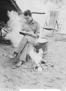 NEW GUINEA, 1942-08. ONE WAY TO COMBAT THE MOSQUITO MENACE IN NEW GUINEA. THIS AUSTRALIAN SOLDIER READS MAIL FROM HOME OVER A FIRE OF SMOKING DRIED COCONUT SHELLS, WHICH GIVES OFF A VERY PUNGENT ..