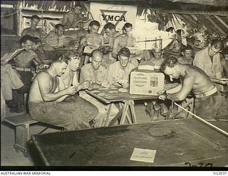 NEW GUINEA. C. 1944. MEMBERS OF AN AIF BAKERY UNIT "SOMEWHERE IN NEW GUINEA" AT A YMCA HUT WITH RAAF PERSONNEL LISTENING TO RACING DESCRIPTIONS RE-BROADCAST FROM THE ABC SHORT-WAVE SERVICE DURING ..