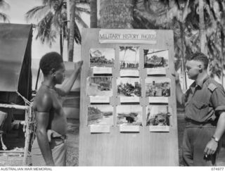 MILILAT, NEW GUINEA 1944-07-26. VX73951 PRIVATE L.S. MORGAN, AUSTRALIAN MILITARY FORCES, AND A NEW GUINEA NATIVE EXAMINING WITH INTEREST THE LATEST PHOTOGRAPHS DISPLAYED ON THE MILITARY HISTORY ..