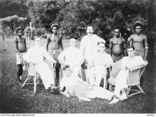 Group portrait of soldiers of the Australian Naval and Military Expeditionary Force (AN&MEF), with their native batmen, enjoying a rest in the garden of their quarters. The young man in front plays ..