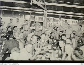 NADZAB, NEW GUINEA. C. 1944-02. MEMBERS OF NO. 24 (VENGEANCE) SQUADRON RAAF HOLD UP POSTERS OF "ADOPTED" TIVOLI GIRL PERFORMERS DURING A MEAL IN THE AIRMEN'S MESS