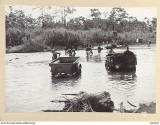 BOUGAINVILLE, 1945-07-11. TROOPS OF 29 INFANTRY BRIGADE WASHING VEHICLES IN THE MOBIAI RIVER WHILE NATIVE CARRIERS TAKE SUPPLIES TO FORWARD COMPANY ON THE MIVO RIVER. HEAVY RAIN HAS MADE THE BUIN ..